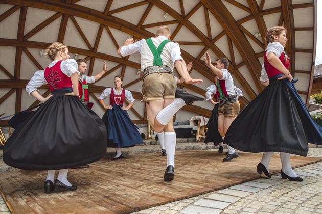 Eine Gruppe von Tänzern in traditioneller bayerischer Tracht tanzt auf einer Bühne. Die Frauen tragen lange schwarze Röcke und die Männer kurze Hosen mit Socken.