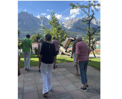Eine Gruppe älterer Menschen spaziert in einer schönen Landschaft mit Bergen im Hintergrund. Der Himmel ist blau und die Umgebung ist grün.