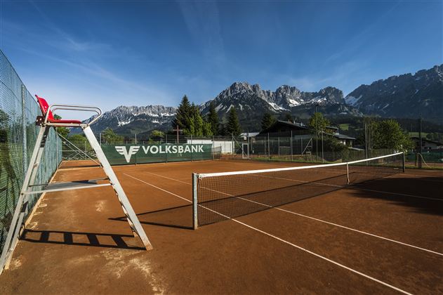 Ein Tennisplatz mit rotem Sand und einer imposanten Bergkulisse im Hintergrund. Die Sonne scheint und der Himmel ist klar.