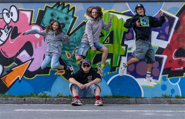 Vier Kinder springen vor einer bunten Graffiti-Wand. Sie haben Spaß und tragen sportliche Kleidung.