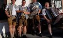 Vier Männer in traditionellen bayerischen Trachten stehen lächelnd zusammen. Sie halten Musikinstrumente wie eine Tuba, ein Akkordeon und Trompeten.
