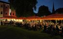 A lively outdoor area with many people sitting at tables and eating. The scene is illuminated in the evening with glowing sun umbrellas.