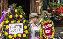 Een vrouw in traditionele klederdracht staat voor een houten huis en draagt twee feestelijk versierde kransen. De kransen zijn met kleurrijke bloemen versierd en dragen de teksten "FLOTTEN LOTTE" en "HÖCKEN HOF".