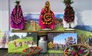 Traditionelle Blumenkränze in bunten Farben stehen auf einem Tisch. Im Hintergrund sind Bilder von der Alm und fröhlichen Menschen mit Kühen zu sehen.