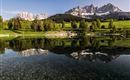 Eine idyllische Landschaft mit einem klaren See und majestätischen Bergen im Hintergrund. Die Bäume spiegeln sich ruhend im Wasser.