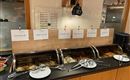 Ein Buffet mit verschiedenen Speisen in warmen Behältern. Darüber hängen Beschilderungen mit den Namen der Gerichte.