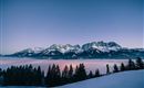Eine beeindruckende Winterlandschaft mit schneebedeckten Bergen und einem hohen Aussichtspunkt. Der Himmel ist in sanften Farben beleuchtet und Nebel zieht durch das Tal.