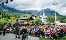 Eine große Zuschauermenge sitzt im Freien vor einer Bühne, während ein Redner spricht. Im Hintergrund sind Berge und eine Kirche zu sehen, was eine malerische Kulisse schafft.