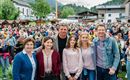 Eine Gruppe von fünf Personen steht lächelnd vor einer großen Menschenmenge. Im Hintergrund sind Berge und Gebäude zu sehen.
