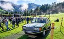 Ein klassisches Auto steht auf einem grünen Feld. Im Hintergrund sind Menschen und Berge zu sehen, und die Atmosphäre wirkt festlich.