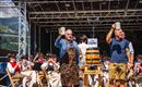 Zwei Männer heben Bierkrüge auf einer Bühne während einer Feier. Im Hintergrund sind Musiker in traditionellen Kostümen zu sehen.