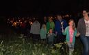 Een groep mensen loopt 's nachts, allemaal met kaarsen in de hand. Op de achtergrond zijn bomen en gras te zien.