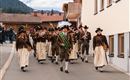 A traditional costume parade with people in festive clothing is walking through a street. In the foreground, a man with a flag leads the group.