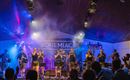 Eine lebhafte Band spielt auf einer Bühne, umgeben von bunten Lichtern und Nebeleffekten. Das Publikum ist begeistert und applaudiert.