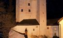Een historische toren bij nacht, omringd door feestelijke verlichting. Veel mensen verzamelen zich om deel te nemen aan een markt.