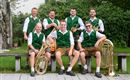 Eine Musikergruppe sitzt im Freien auf einer Bank. Sie tragen grüne Westen und spielen verschiedene Blasinstrumente.