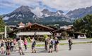 Een groep mensen staat voor een rustiek gebouw in de Alpen. De indrukwekkende bergen zijn op de achtergrond zichtbaar, omgeven door wolken.