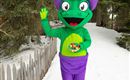 Een kleurrijk mascotte staat in de sneeuw en zwaait. Het draagt een paarse hoed en houdt een kom met kleurrijke snoepjes vast.