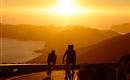 Zwei Radfahrer fahren einen kurvenreichen Weg entlang einer Küste bei Sonnenuntergang. Die Szenerie ist von warmem Licht und sanften Hügeln umgeben.