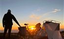 To cyklister kører ad en snoet vej langs en kyst ved solnedgang. Scenen er omgivet af varmt lys og bløde bakker.