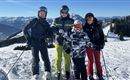 Eine Gruppe von vier Skifahrern steht auf einem schneebedeckten Hang. Im Hintergrund sind Berge und ein klarer Himmel zu sehen.