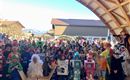 A large group of children in colorful costumes gathers under a wooden roof. In the background, mountains and a clear sky are visible.