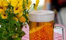 Sklenice piva s pěnivou korunou stojí vedle kytice žlutých květin. Stůl má červeno-bílo kostkovaný ubrus.
