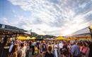 Ein lebendiger Markt mit vielen Menschen und bunten Ständen. Die Sonne scheint am Himmel und schafft eine fröhliche Atmosphäre.