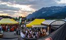 Eine belebte Veranstaltung mit vielen Menschen und bunten Zelten. Im Hintergrund sind imposante Berge und ein schöner Sonnenuntergang zu sehen.
