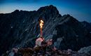 Eine Fackel brennt auf einem felsigen Berggipfel in der Dämmerung. Die beeindruckende Berglandschaft bildet den Hintergrund.