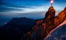 A picturesque mountain panorama at twilight with a burning candle on a rock. The scene shows snow-covered mountains and a clear sky in the background.