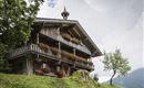 A traditional wooden house with a balcony and colorful flowers. It stands in a green landscape under a cloudy sky.
