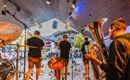 Eine Band spielt auf einer Bühne vor einem Publikum. Im Vordergrund sind Musiker mit Blasinstrumenten zu sehen.