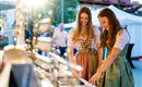 Zwei Frauen in traditionellen Kleidern schauen sich Schmuck auf einem Markt an. Die Sonne scheint, und im Hintergrund sind andere Besucher zu sehen.