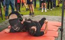 Zwei Kämpfer liegen auf roten Matten und sind im Wettkampf miteinander beschäftigt. Eine Gruppe von Zuschauern beobachtet das Geschehen im Hintergrund.