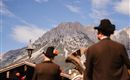 Two men in traditional costumes stand in the foreground and look at majestic mountains. The sky is blue with some clouds.
