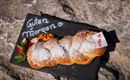 Ein frisch gebackenes, geflochtenes Brot mit Puderzucker auf einem schwarzen Steinuntergrund. Daneben steht "Guten Morgen" auf dem Stein, dekoriert mit bunten Blumen.