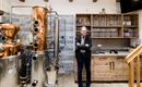 A modern distillery with a copper kettle and wooden shelves. A man stands relaxed next to the equipment.