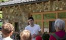 Ein junger Mann spricht zu einer Gruppe von Menschen vor einem Steinhaus. Die Zuhörer sind aufmerksam und scheinen interessiert zu sein.