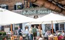 Eine belebte Terrasse eines Gasthauses mit Gästen unter Sonnenschirmen. Im Hintergrund ist das Schild "Gasthof Stangl" sichtbar.