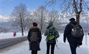 Drie personen wandelen op een sneeuwbedekte weg. Op de achtergrond zijn besneeuwde bergen en bomen te zien.