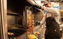 Ein Koch steht am Grill und bereitet Fleisch auf einem Spieß zu. Im Vordergrund sind Pommes frites und ein Burger zu sehen.
