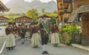 Eine festliche Parade mit einer Gruppe von Menschen in traditioneller Tracht. Im Hintergrund sind malerische Berge und Holzhäuser zu sehen.