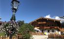 Ein traditionelles Holzhaus mit Balkon und schönen Blumen vor der Tür. Im Hintergrund ist ein blauer Himmel mit einigen Wolken zu sehen.