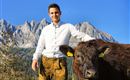 Ein junger Mann steht neben einem Rind in der Natur. Im Hintergrund sind beeindruckende Berge und ein blauer Himmel zu sehen.