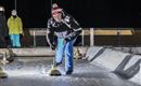 Eine Person spielt Curling auf einer Eisbahn. Im Hintergrund sind Zuschauer in winterlicher Kleidung zu sehen.