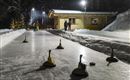 Eine Eisbahn, beleuchtet von Laternen, mit fünf Holzstöcken auf dem Eis. Im Hintergrund stehen mehrere Personen und eine kleine Hütte.