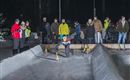 Eine Gruppe von Menschen beobachtet ein Kind, das auf einer Eisbahn spielt. Die Umgebung ist winterlich und es ist dunkel, mit nur wenigen Lichtern sichtbar.