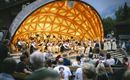 Ein Orchester spielt in einem einzigartigen Pavillon vor einem Publikum. Menschen sitzen auf Bänken und genießen das Konzert.