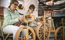 Zwei Frauen sitzen an Spinnrädern und arbeiten mit Wolle. Sie tragen kariert gestreifte Blusen und konzentrieren sich auf ihre Handarbeit.